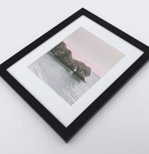 A big dark gray rock popping out of the ocean in a light ping sunrise on a photo print in a black frame